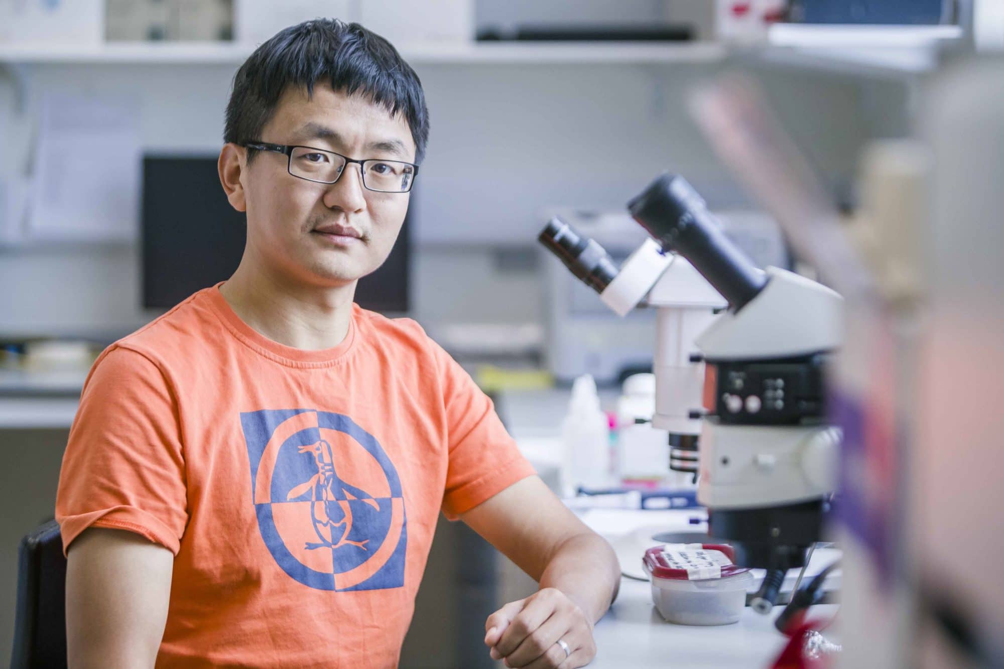Jing Yu - a man wearing an orange t shirt facing the camera. There is a microscope to his left on a desk.
