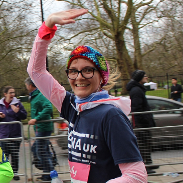 Image shows a runner in a blue Retina UK t-shirt. She is waving as she runs by.
