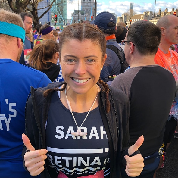 Image shows a runner in a blue running vest with 'Sarah - Retina UK' on the front in white font. Sarah is smiling and has her 'thumbs up'.