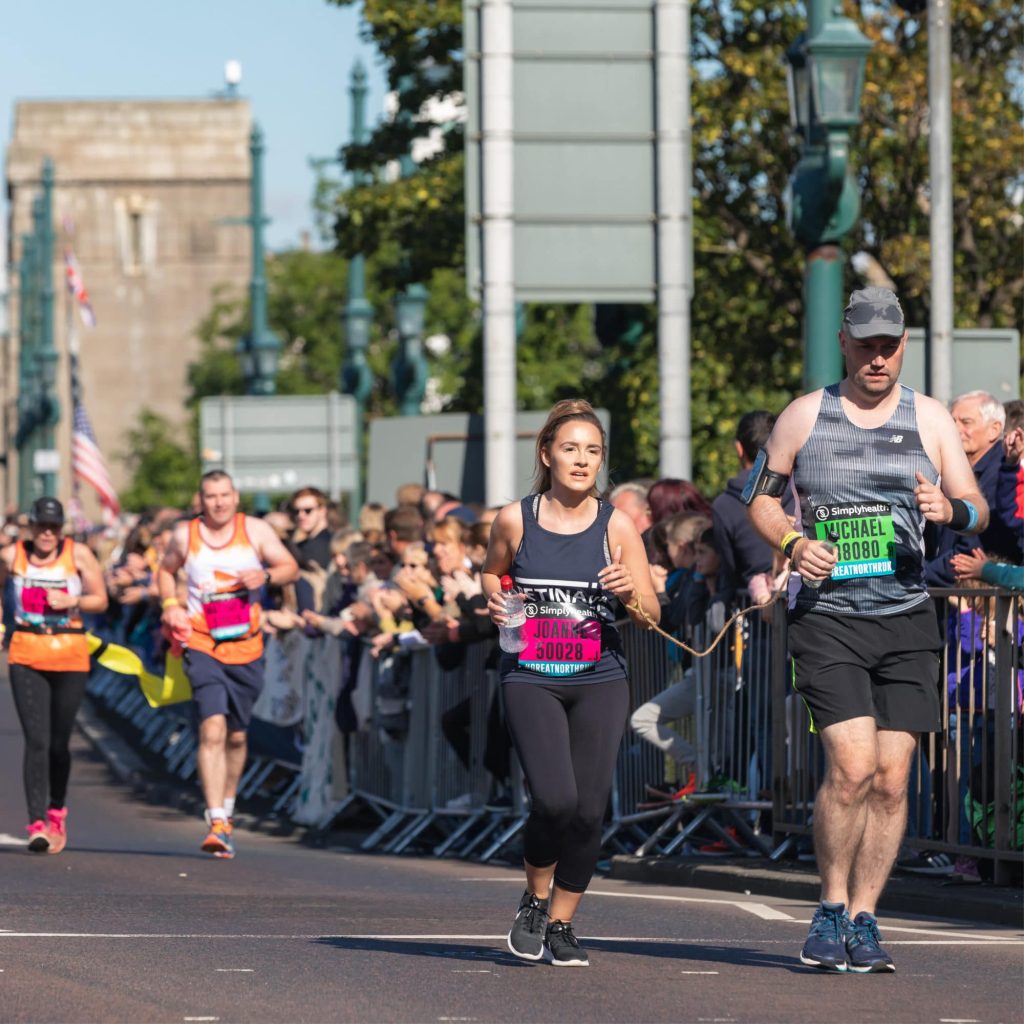 Image shows a runner in a blue Retina UK running vest with her guide runner to the right. Crowds line the road as they run by.