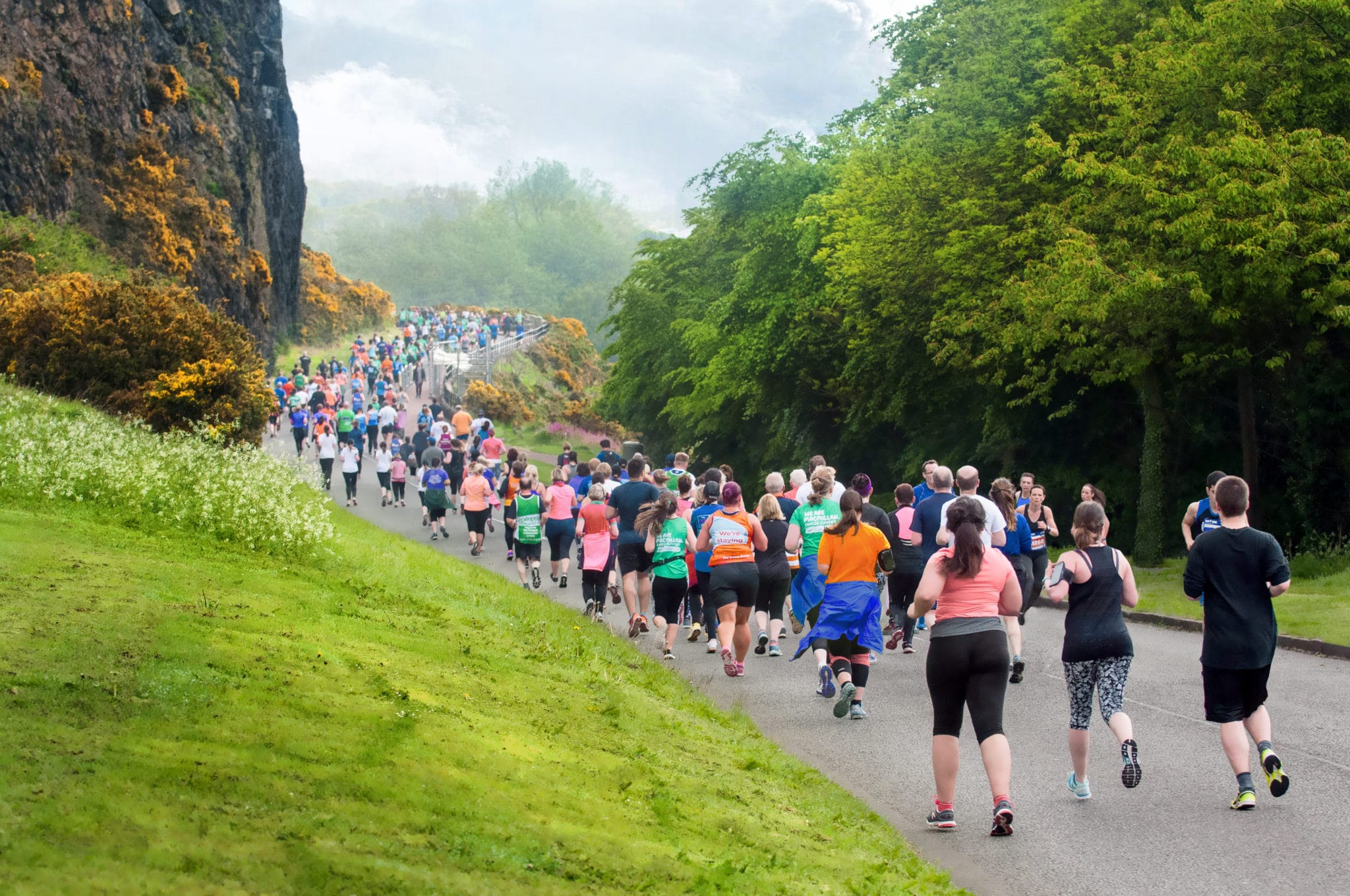 Image shows hundreds of runners running down a path in Holyrood Park. Yellow flowering gorse contrasts against the dark rock cliffs.