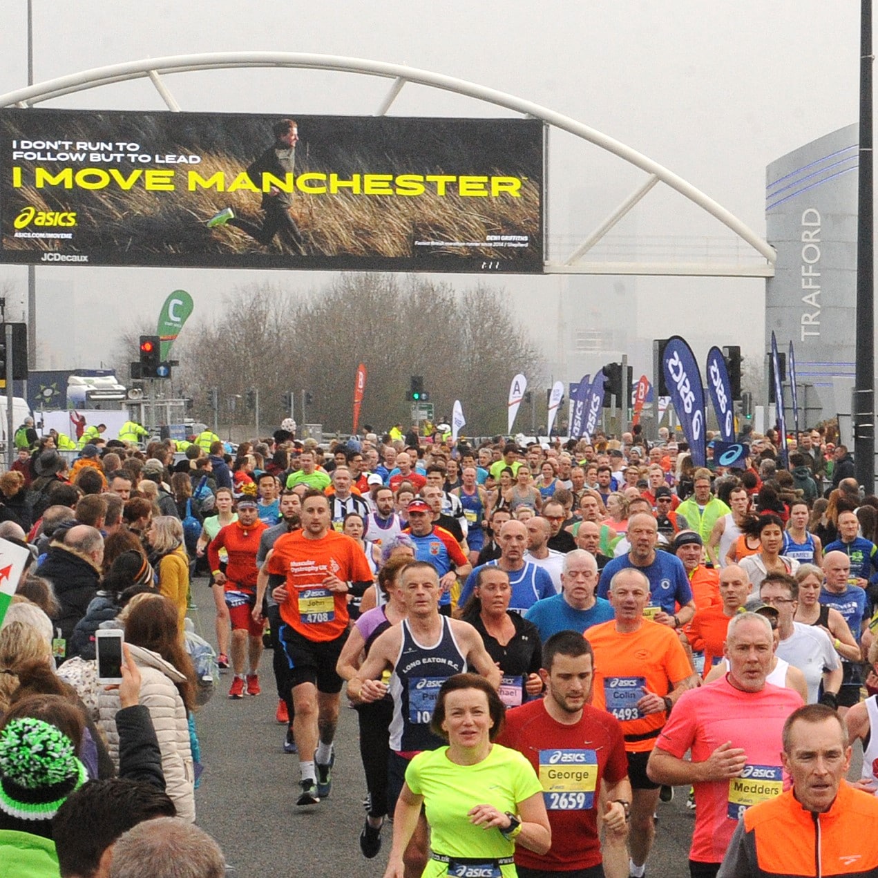 Image shows runners running down a road with a sign above that reads 'I Move Manchester'