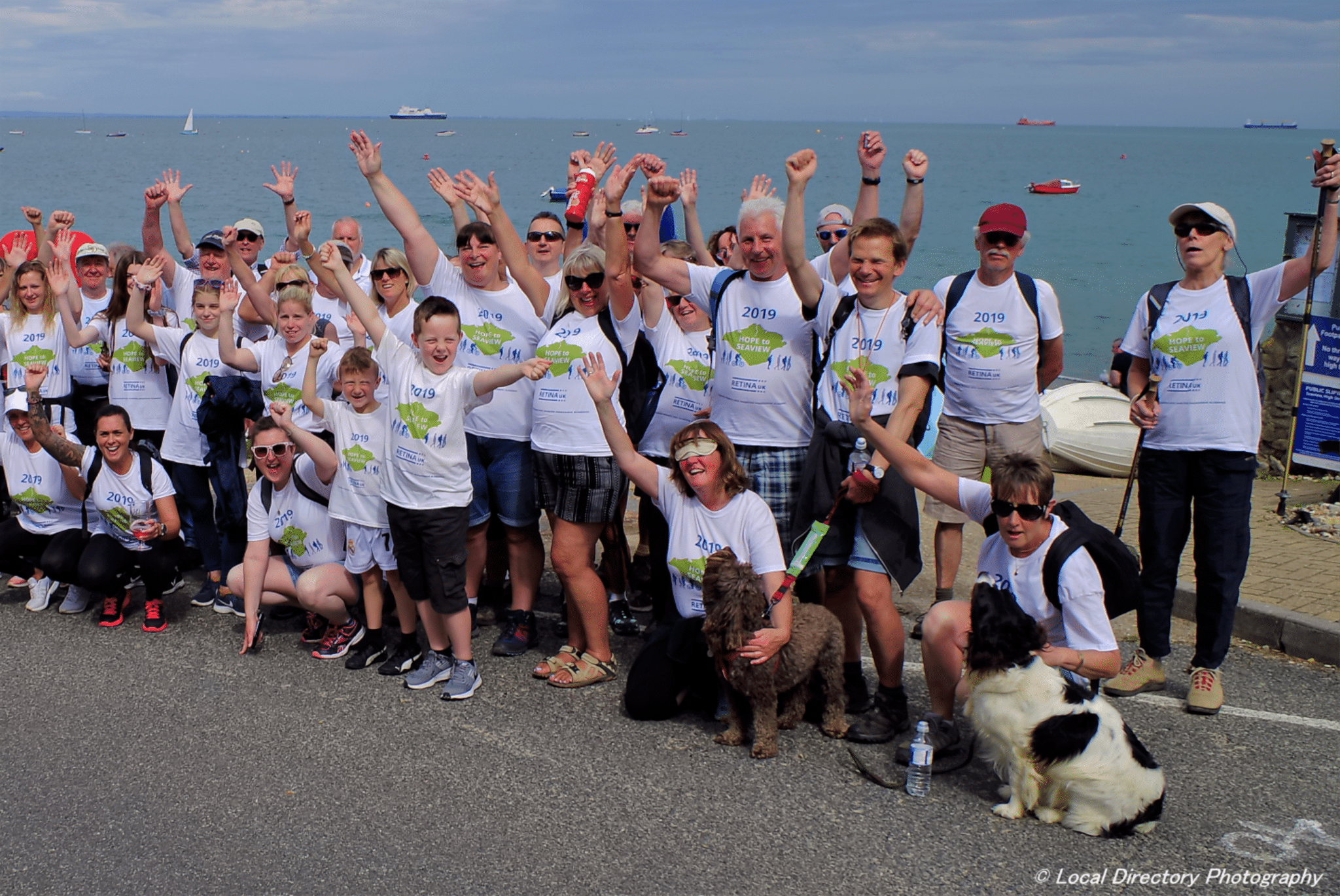 Image shows a crowd of supporters grouped together and cheering. They are wearing white Hope to Seaview 2019 t-shirts. The beach and sea can be seen in the background.