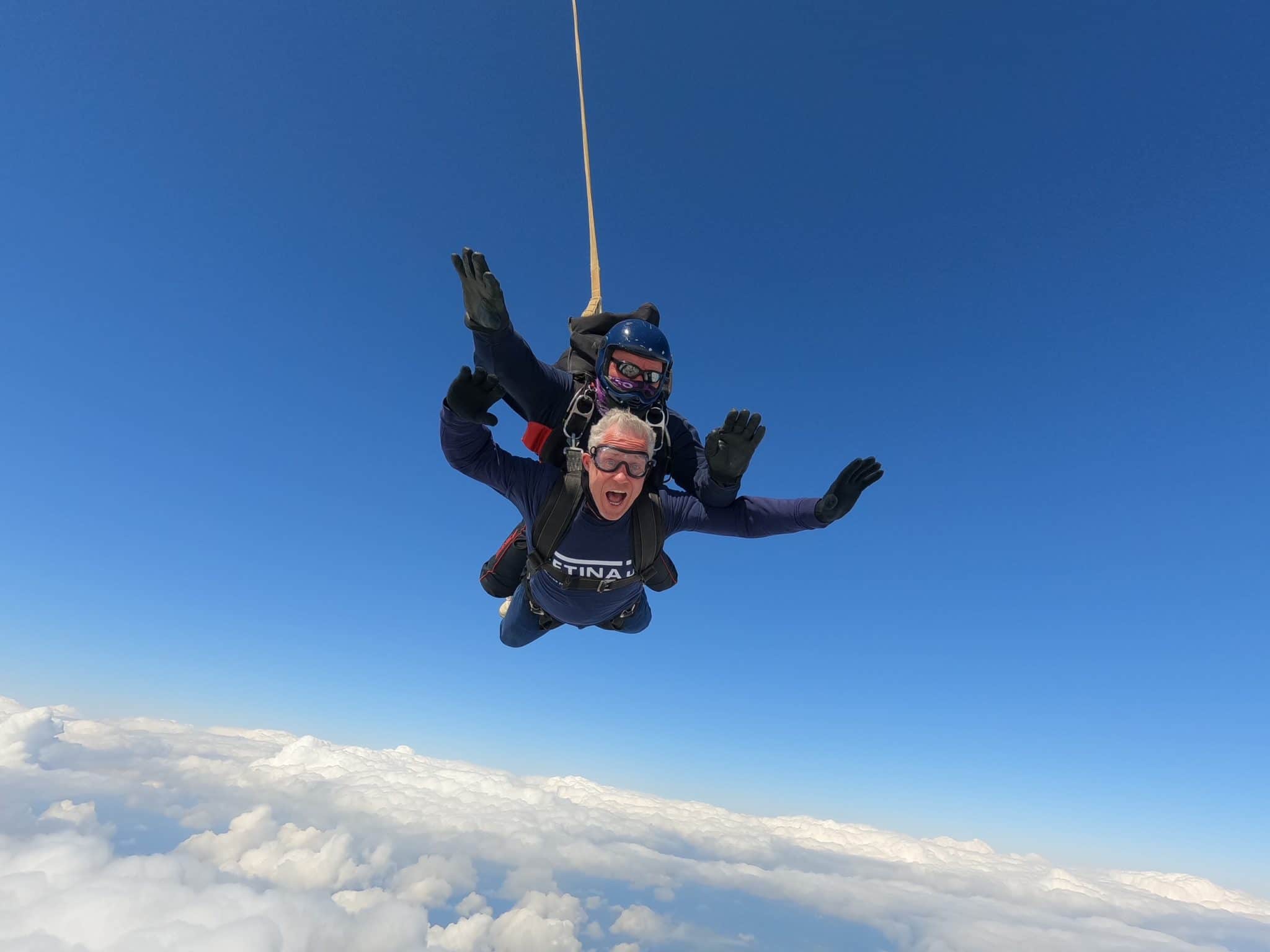 Image shows two people in a tandem skydive - blue sky and clouds are visible.