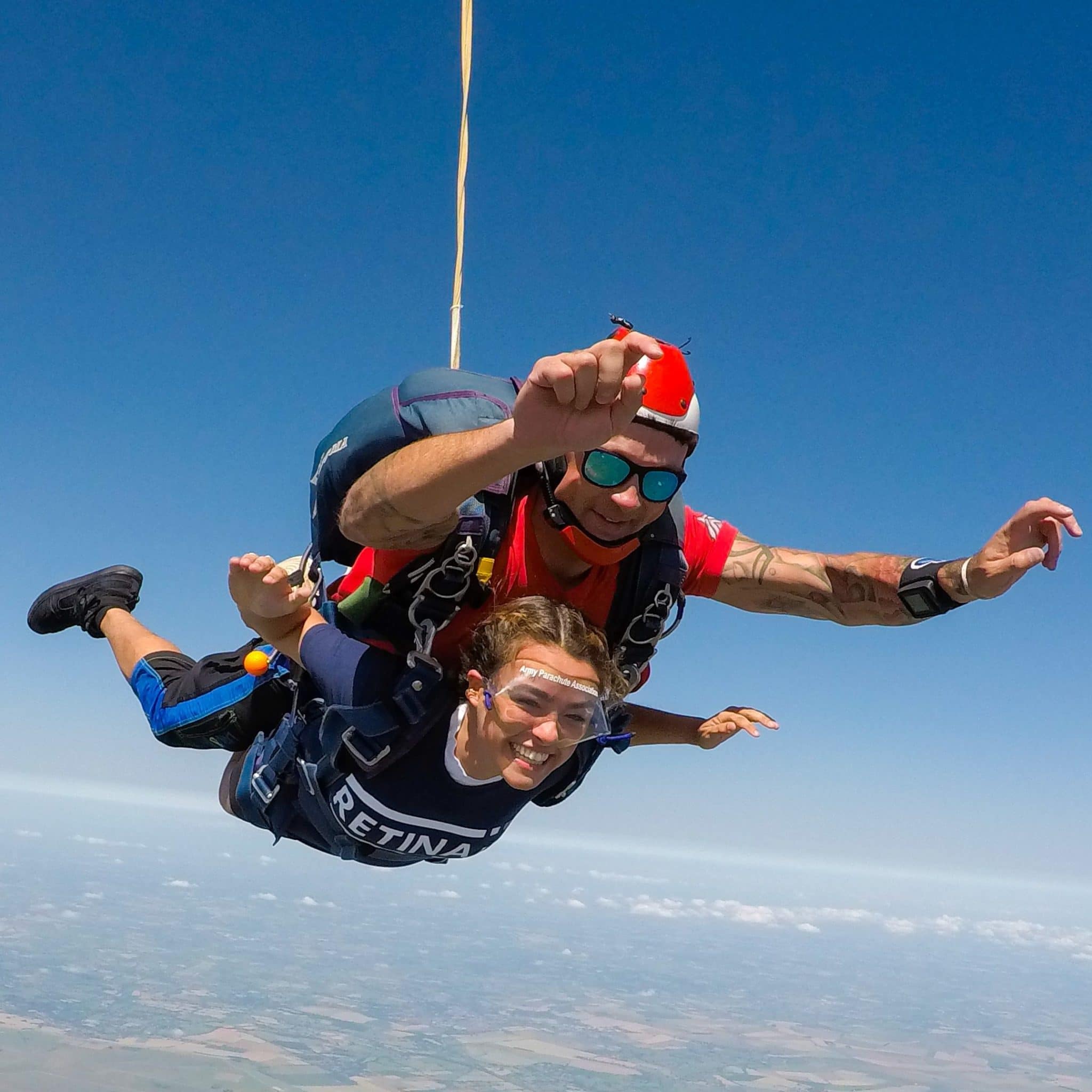 Image shows a Retina UK supporter with a skydive instructor freefalling in tandem. A blue sky is visible with clouds and the ground below.