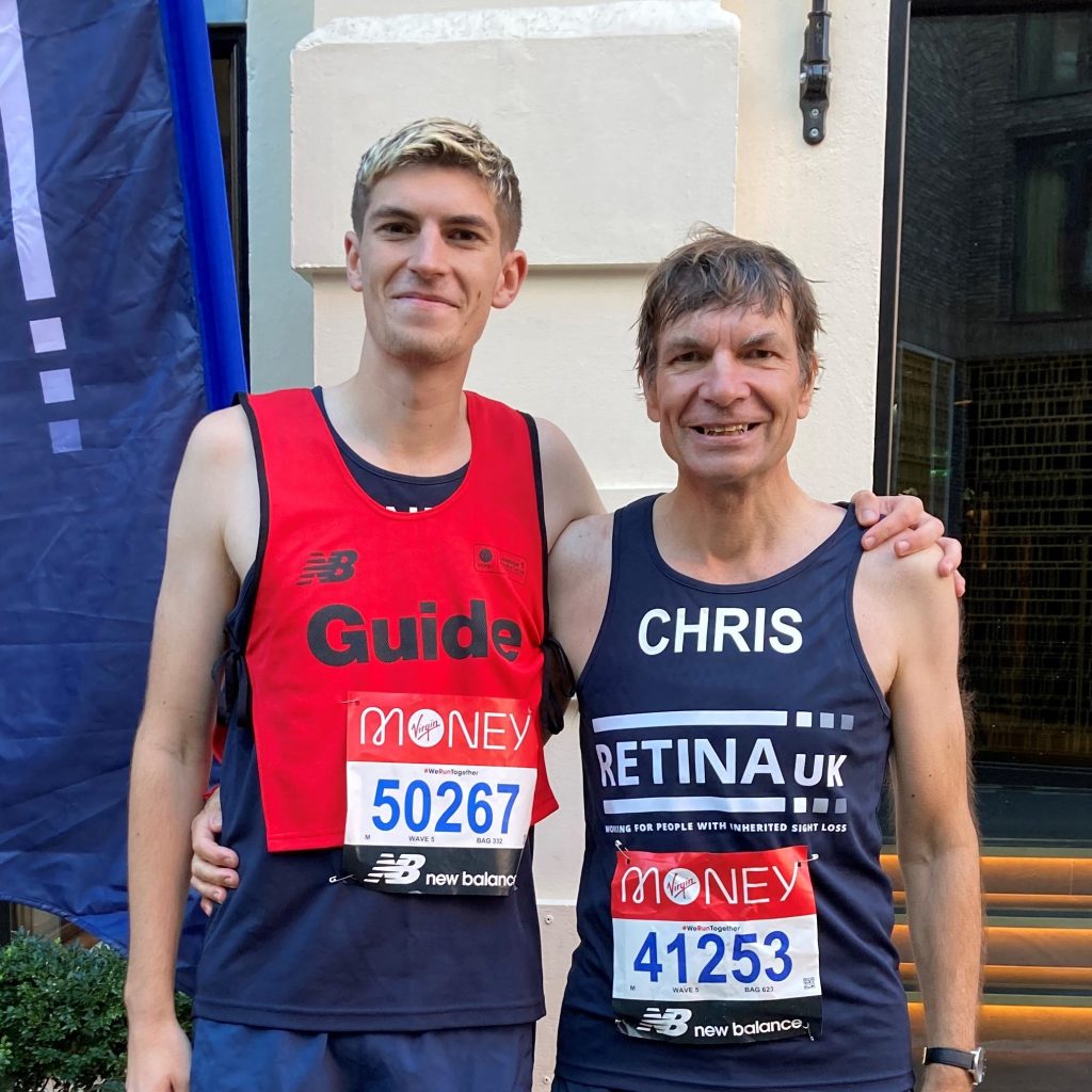 Image shows a guide runner in a red guide bib stood next to a runner in a blue Retina UK running vest. Both are smiling.