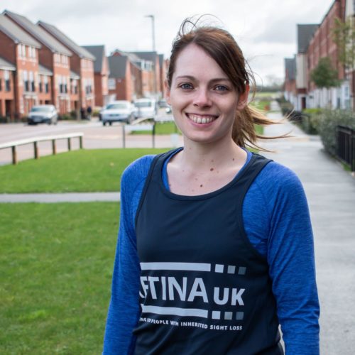 Image shows a runner wearing a blue top and blue Retina UK running vest. She is smiling and is stood in a residential street with houses in the background