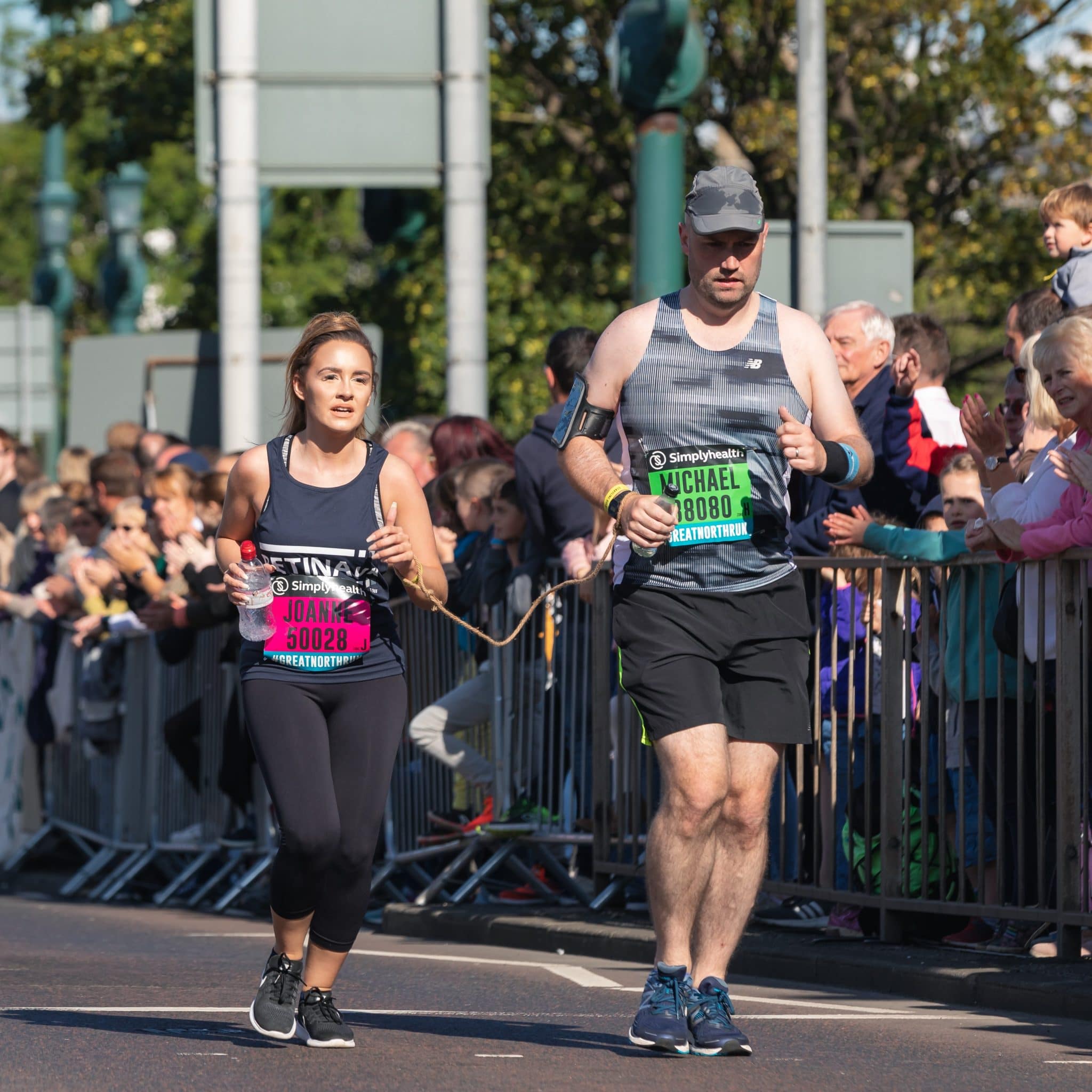 Image shows a runner in a blue Retina UK running vest with her guide runner to the right. Crowds line the road as they run by.