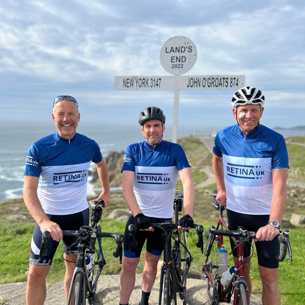 Image shows three cyclists, each wearing blue and white Retina UK cycling jerseys, stood in front of the Land End sign.