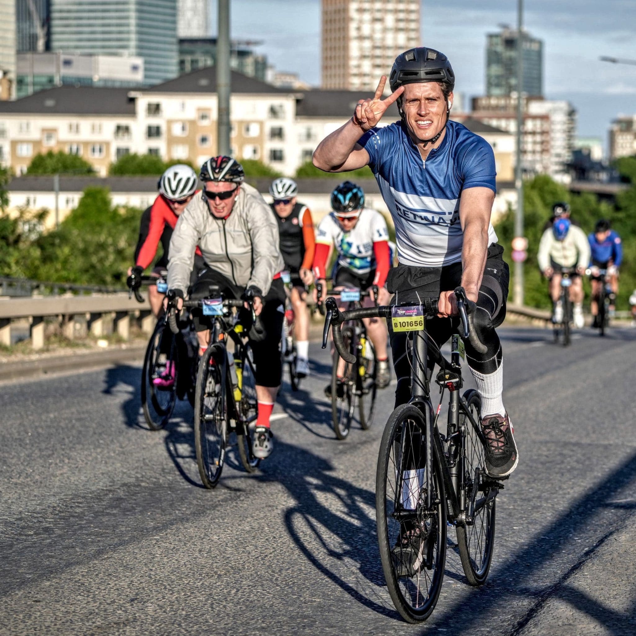 Image shows a cyclist in a blue cycling jersey with 'Retina UK' on the front. He is cycling on a closed road with other cyclists and London buildings in the background.