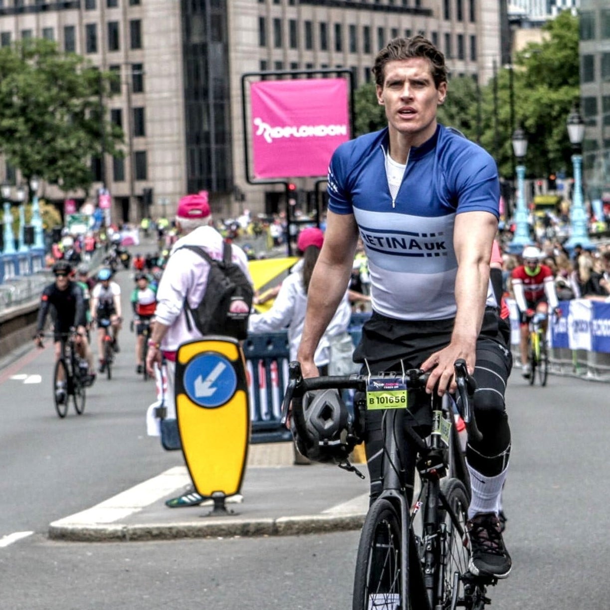 Image shows a cyclist in a blue cycling jersey with 'Retina UK' on the front. He is cycling on a closed road with other cyclists and a RideLondon sign in the background.