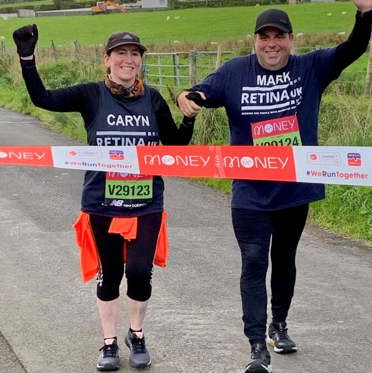 Image shows two walkers completing the Virtual London Marathon. They are waving as they are about to cross a red and white paper finish line.