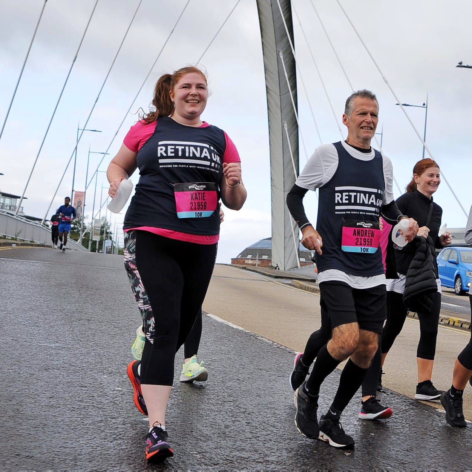 Image shows two runners running along a path. Both are wearing blue Retina UK running vests and pink 10K running numbers.