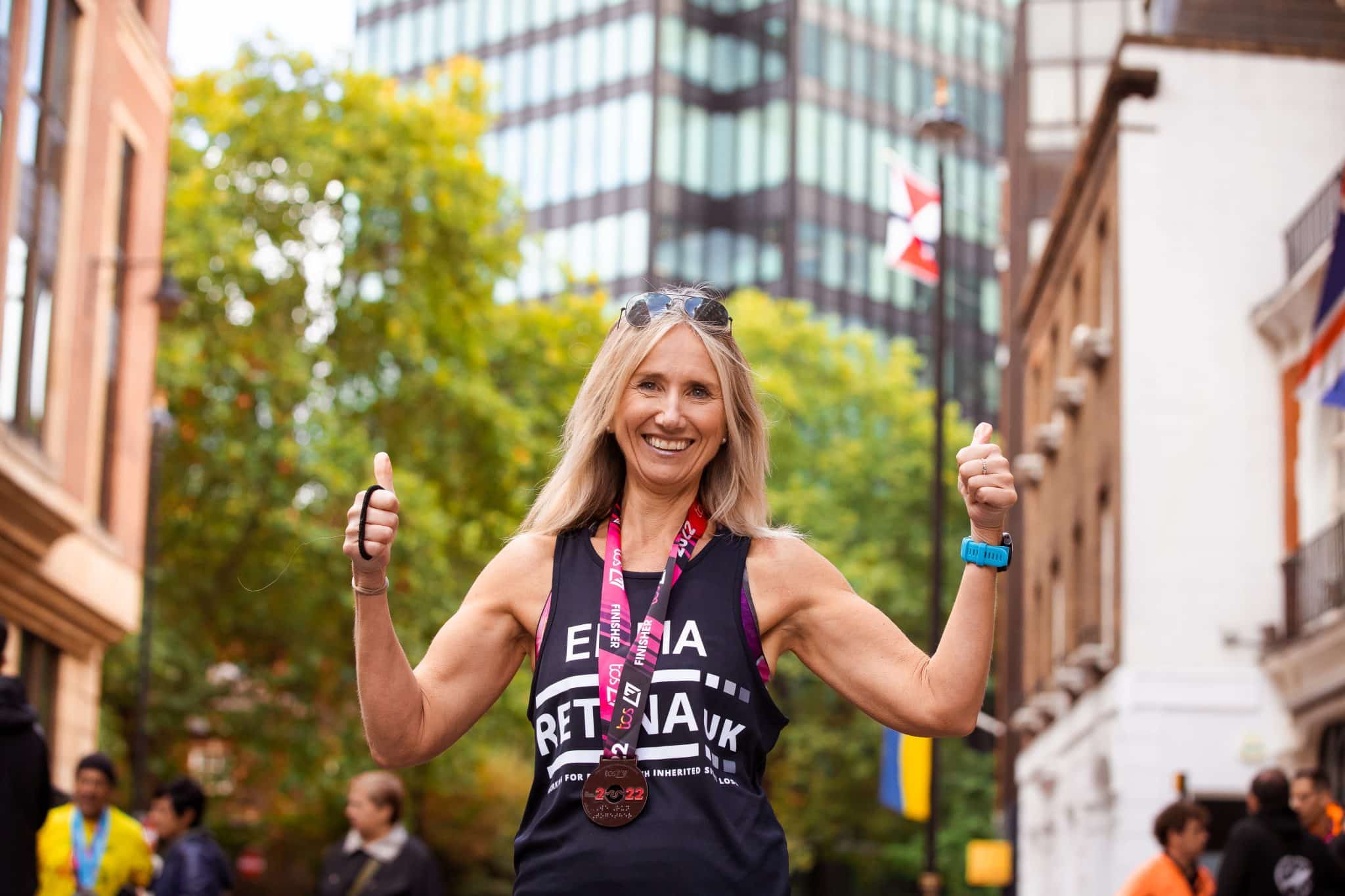 Image shows a runner stood smiling with her thumbs up. She is wearing a blue Retina UK running vest with the 'Retina UK' logo in white on the front. She is also wearing a 2022 London Marathon medal.