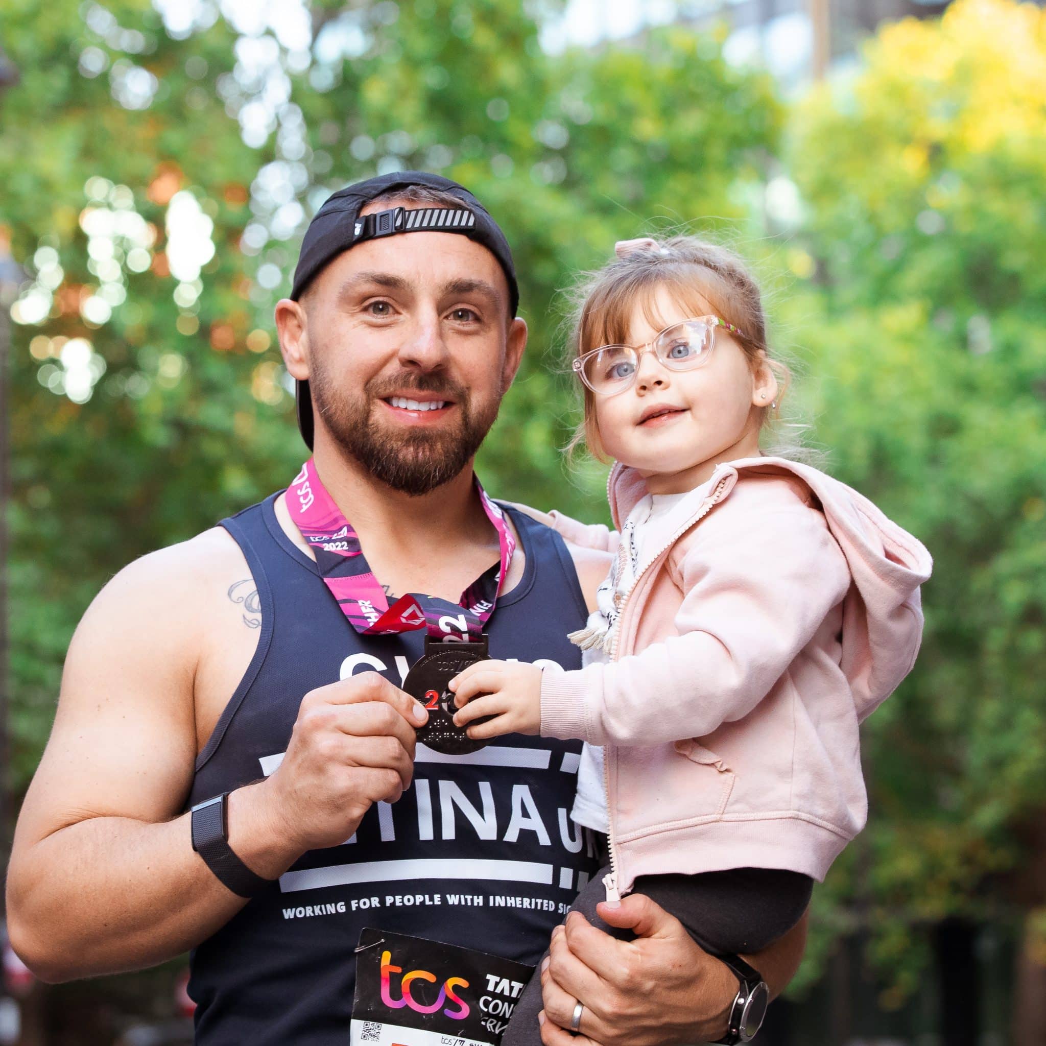 Image shows a man wearing a blue Retina UK running vest and a London Marathon medal. He is holding his young daughter.
