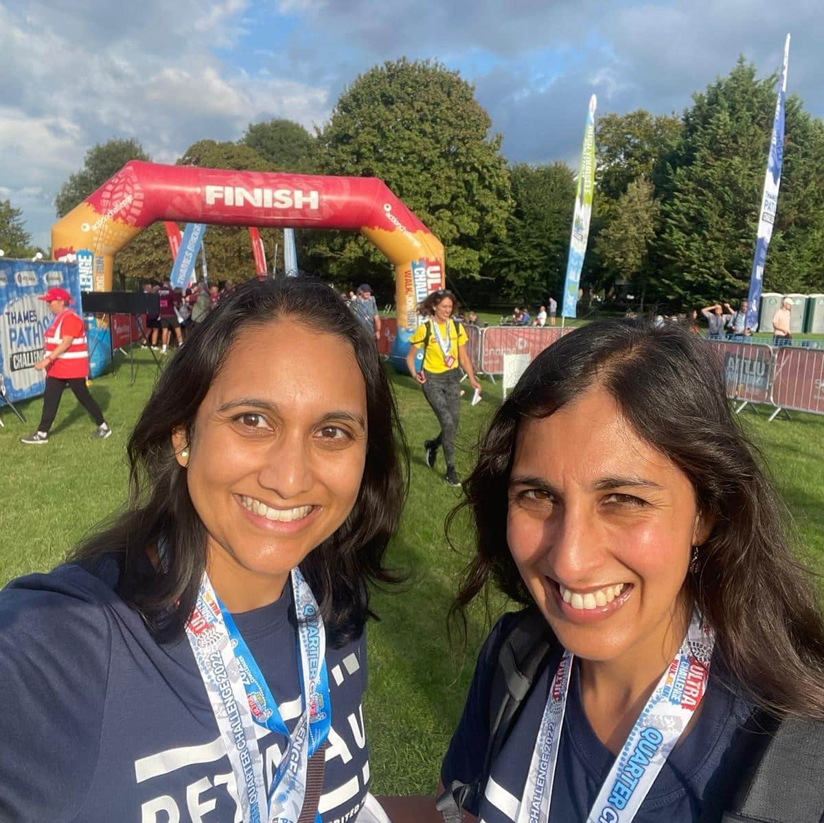 Image shows two women stood next to each other smiling. Both are wearing blue Retina UK t-shirts and their trek medals. An inflatable finish line is visible in the background.