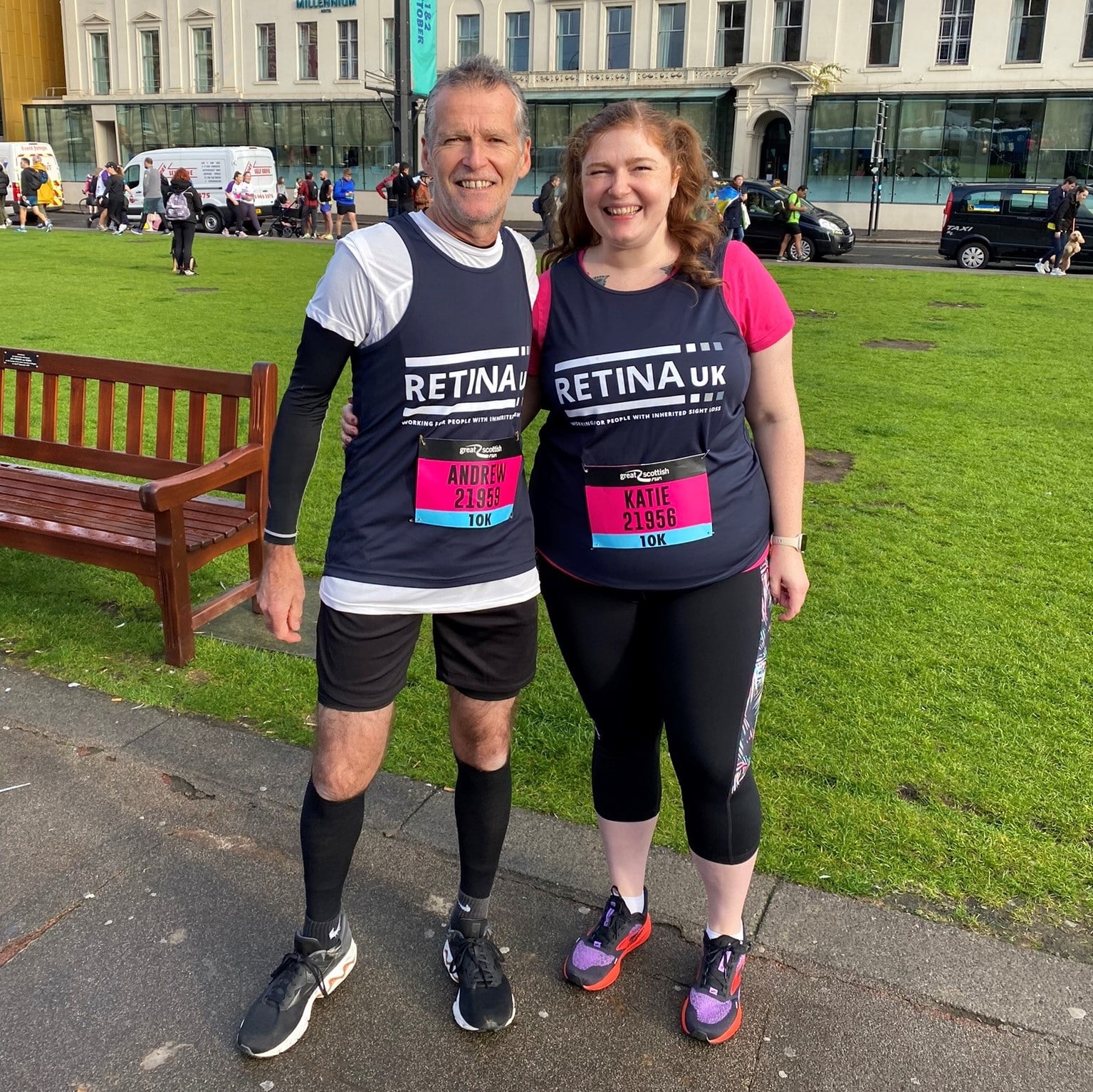 Image shows two runners stood side by side smiling. Both are running 10k running numbers and blue Retina UK tops.