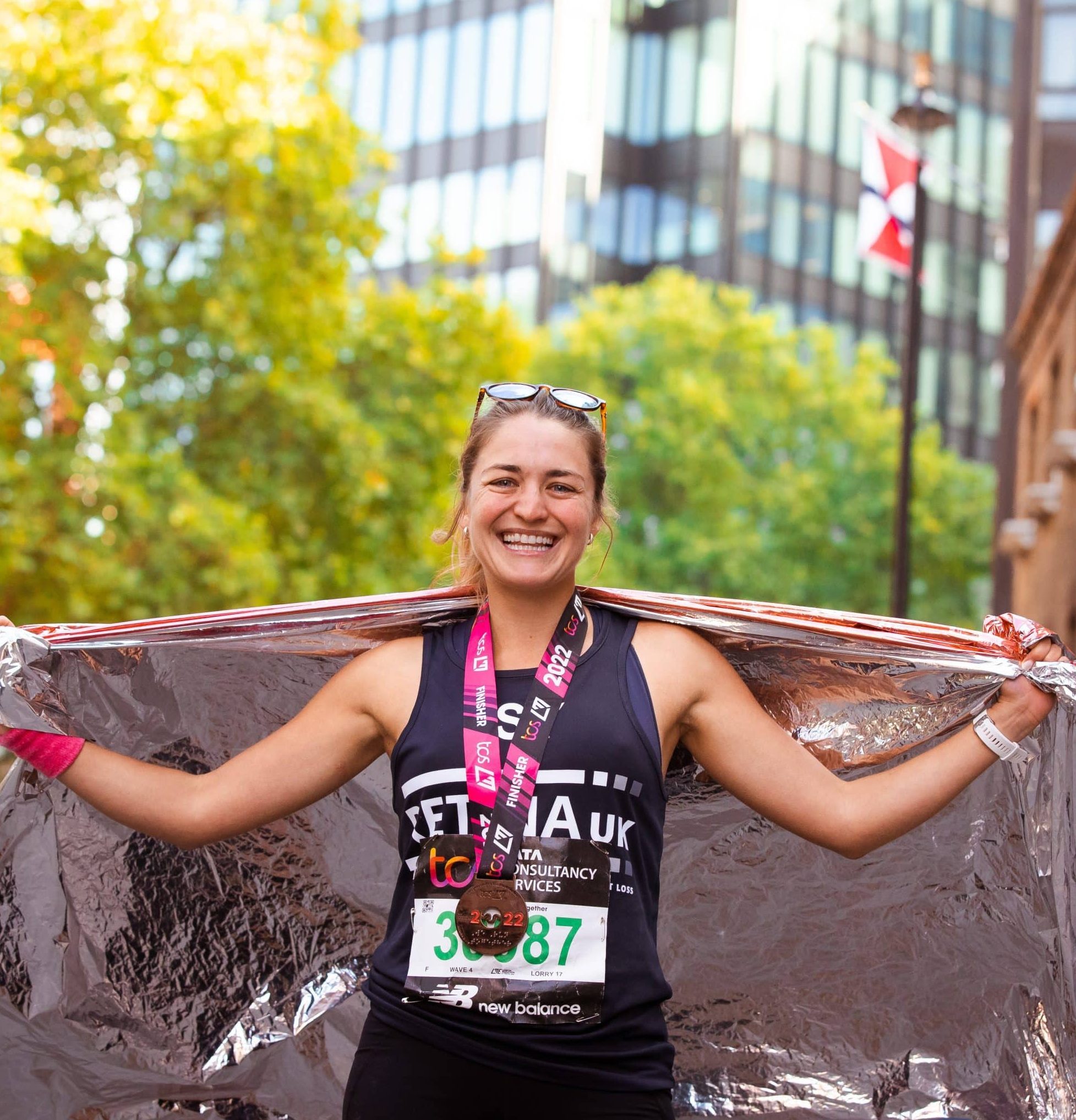 Image shows a runner stood smiling with her arms outstretched . She is wearing a blue Retina UK running vest and a London Marathon medal around her neck.