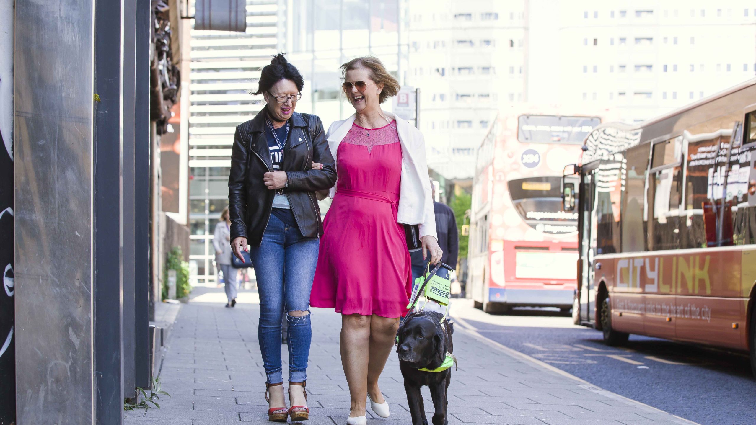 Two women walking along the street. One is wearing a pink dress and white cardigan and is holding a guide dog hardness (the dog is walking to her side). The other woman is wearing jeans and a leather jacket.
