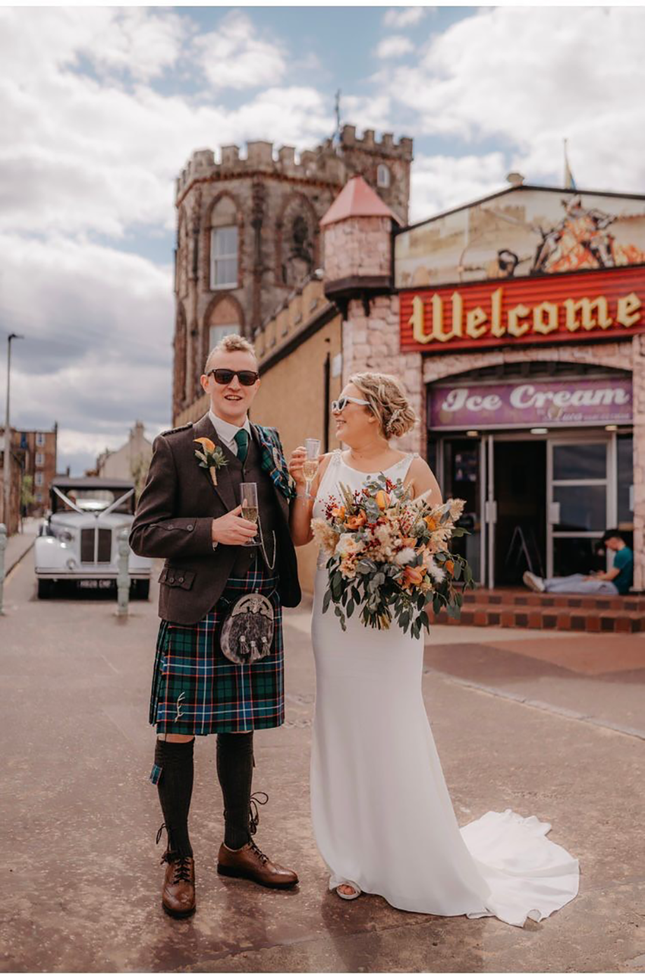 A man in a kilt and a woman in a wedding dress. They are both holding glasses of champagne