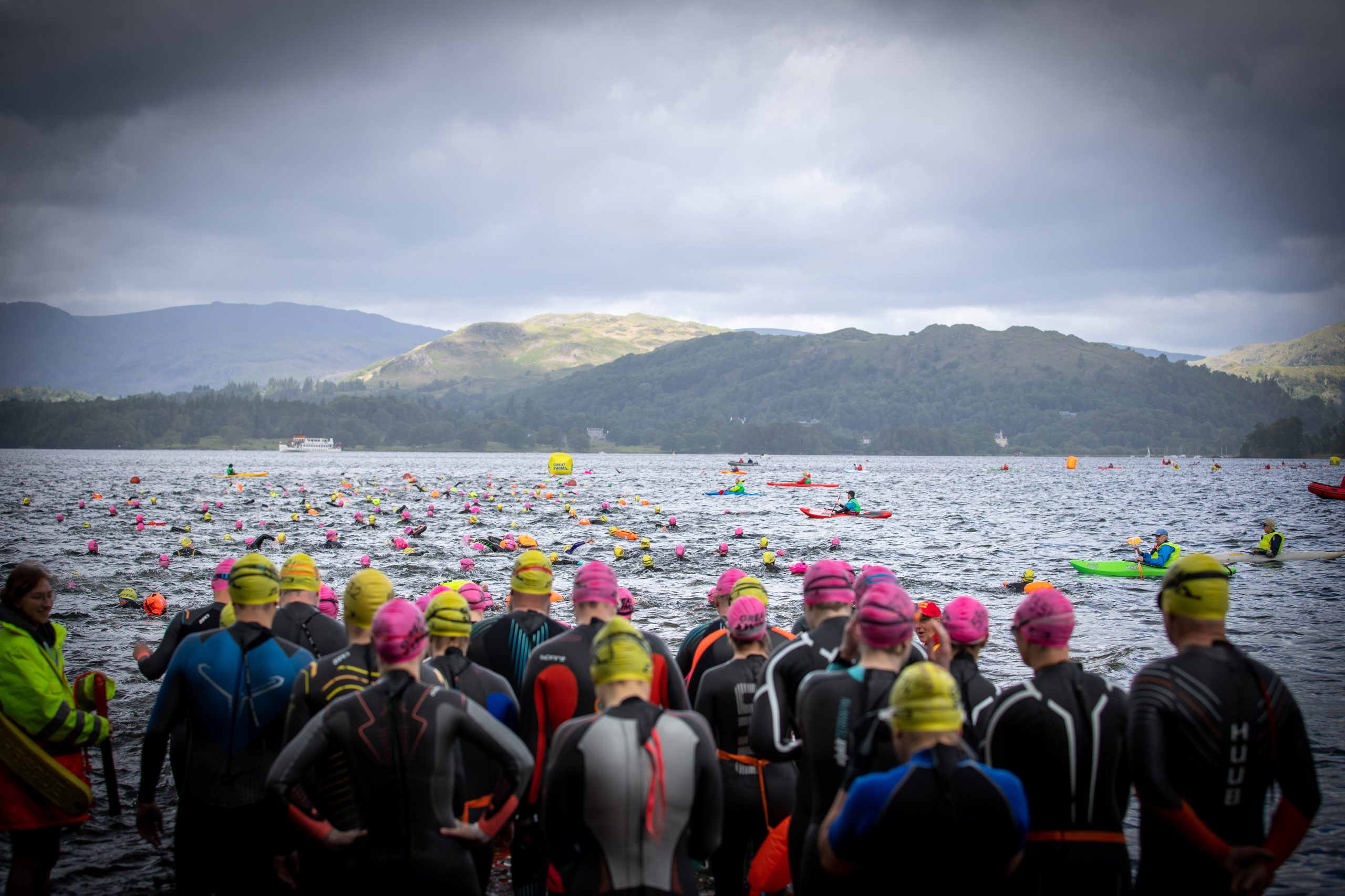 A group of people all wearing brightly coloured swimming caps standing in the foreground with a lake in the background and hills beyond