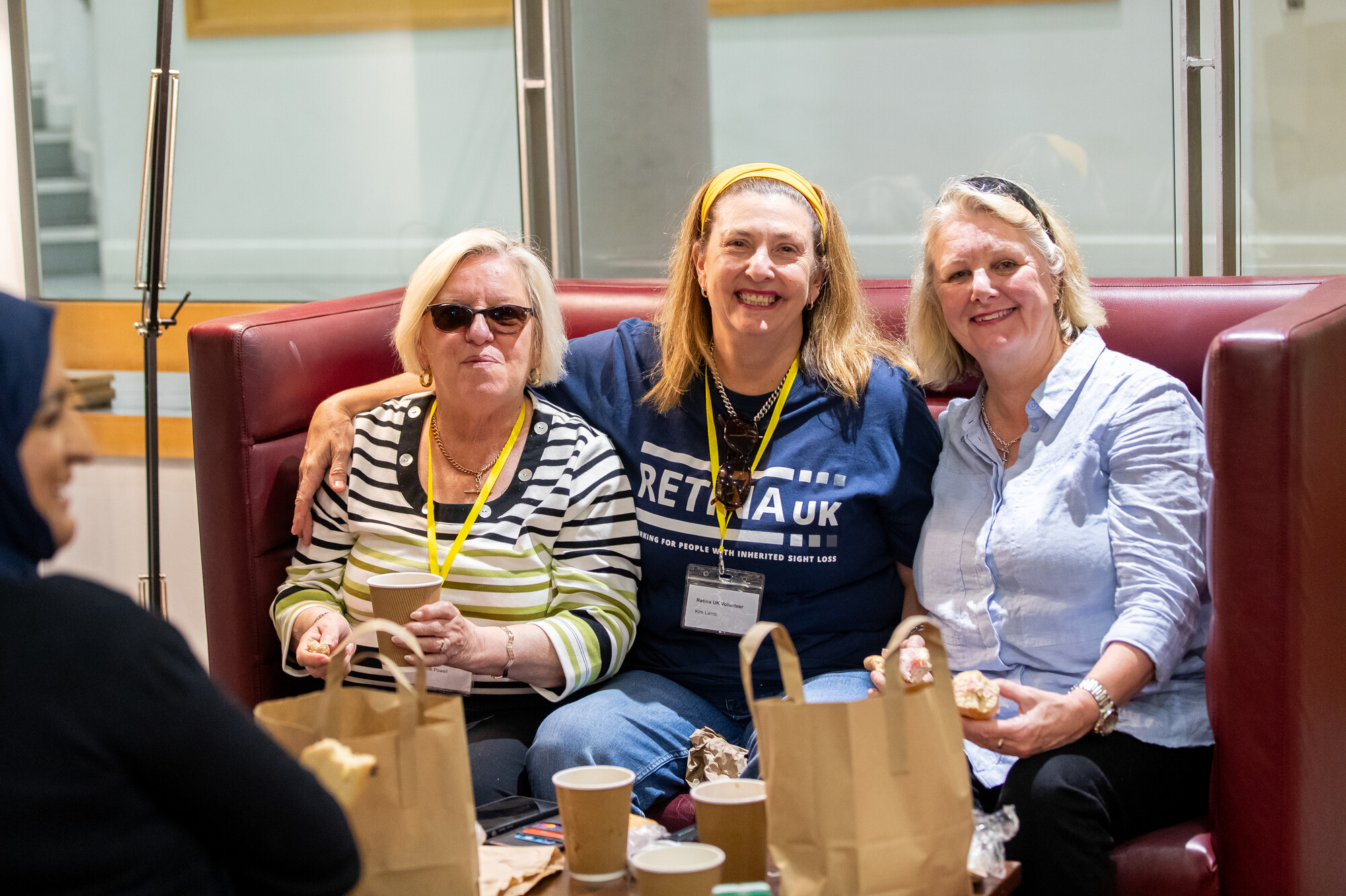 Three ladies sitting together on a sofa. They are all smiling at the camera. The lady in the centre is wearing a Retina UK tshirt.