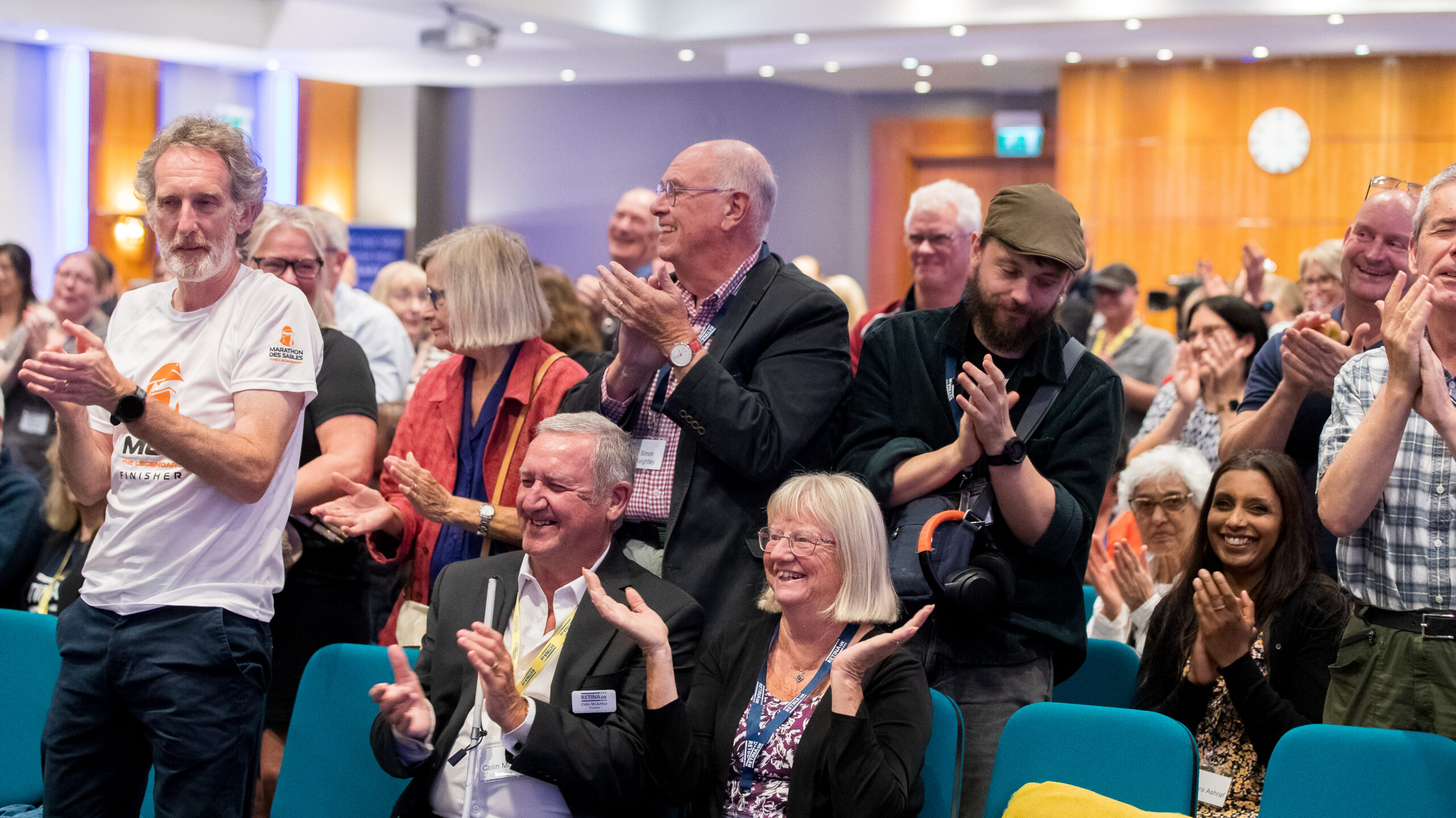 Image shows audience members from 2025 annual conference. Audience are sat and stood applauding and smiling at the stage.