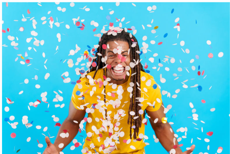 A man wearing a yellow tshirt against a light blue background. He is surrounded by confetti