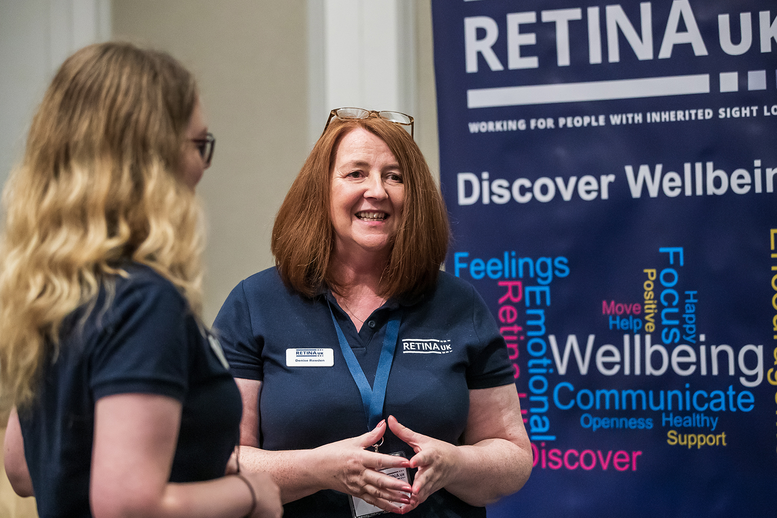 Two women, one facing the camera and one seen from the side. They are both wearing blue short sleeved polo shirts. In the background there is the Retina UK logo and the words Discover Wellbeing