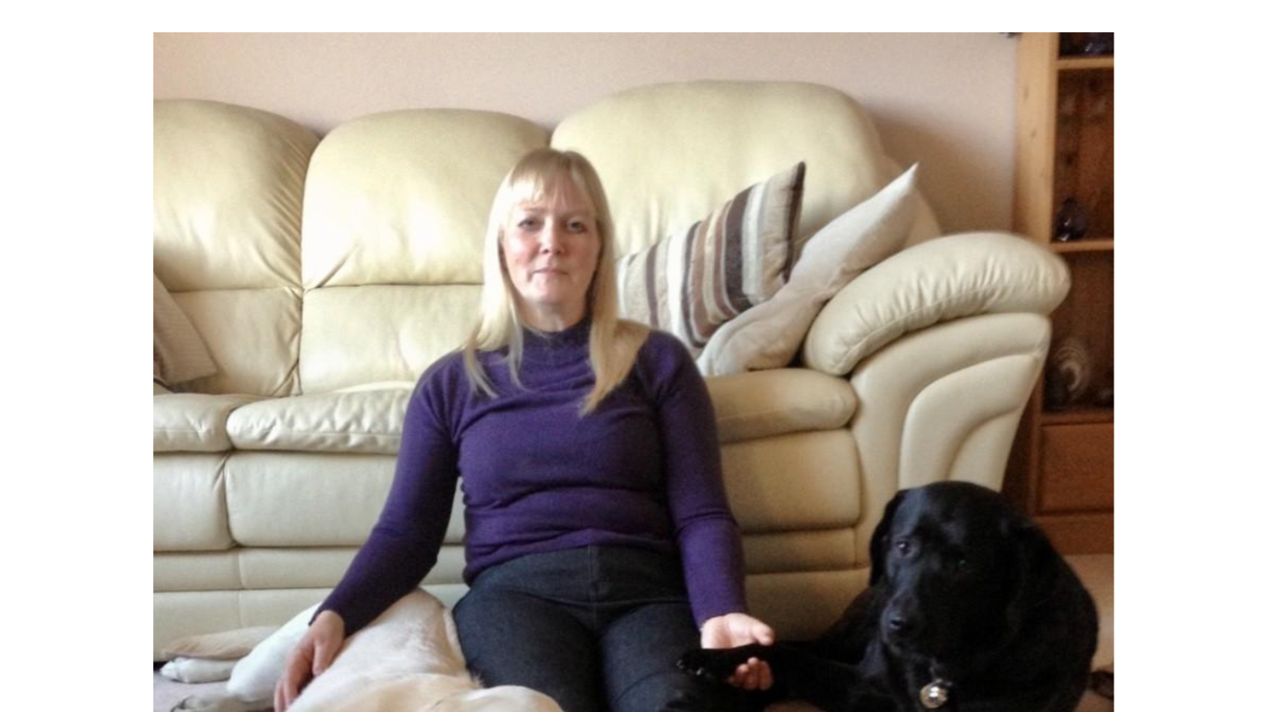 Catherine - a woman will shoulder length fair hair. She is seated on the floor with a yellow labrador on one side of her and a black labrador on the other.