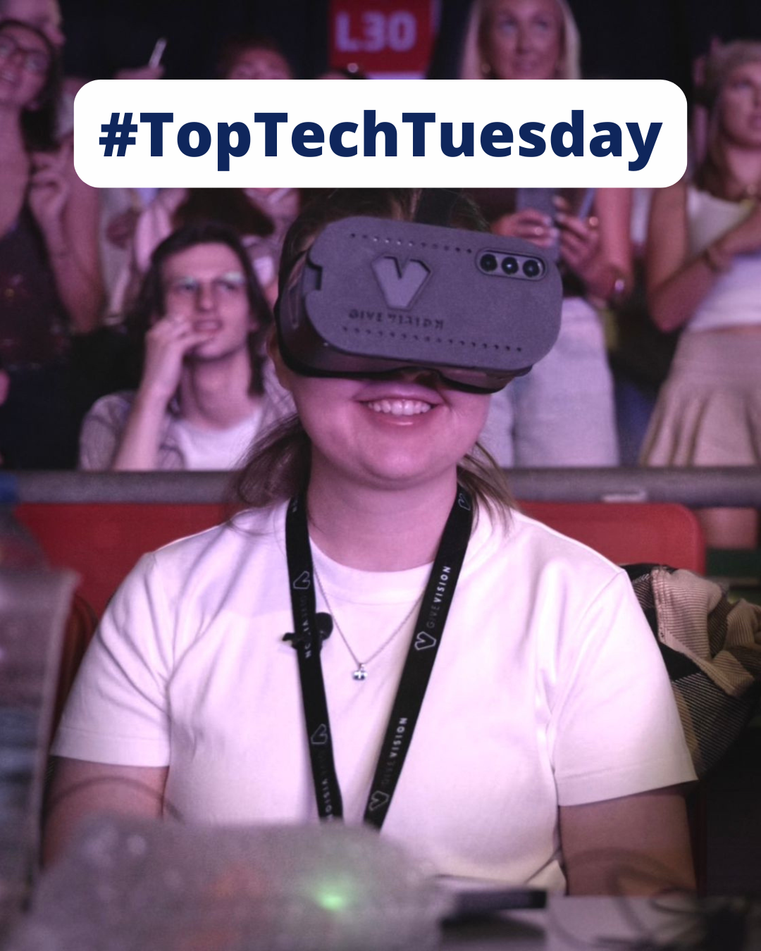 picture of Eve sat watching a concert with the GiveVision headset across her eyes. She is smiling and wearing a white shirt with a lanyard around her neck. Behind her are other people watching the concert. A banner is at the top of the screen with the blue text 