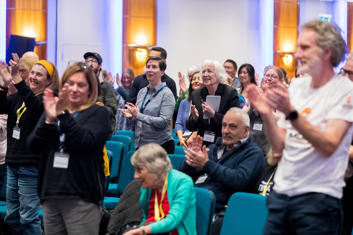 A large group of people standing clapping their hands. They are all facing towards a stage