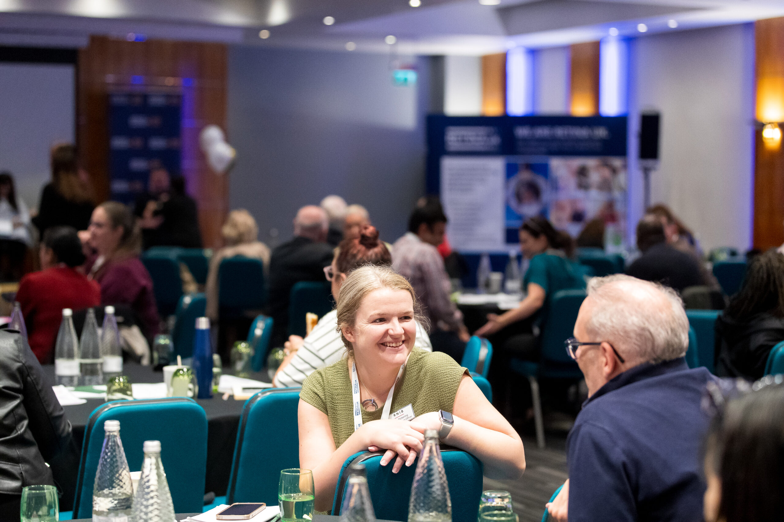 Picture from Retina UK Professionals' Conference 2025. Shows the conference room with round tables and chairs and Retina UK banners. Two people are sitting on the chairs and smiling at each other.