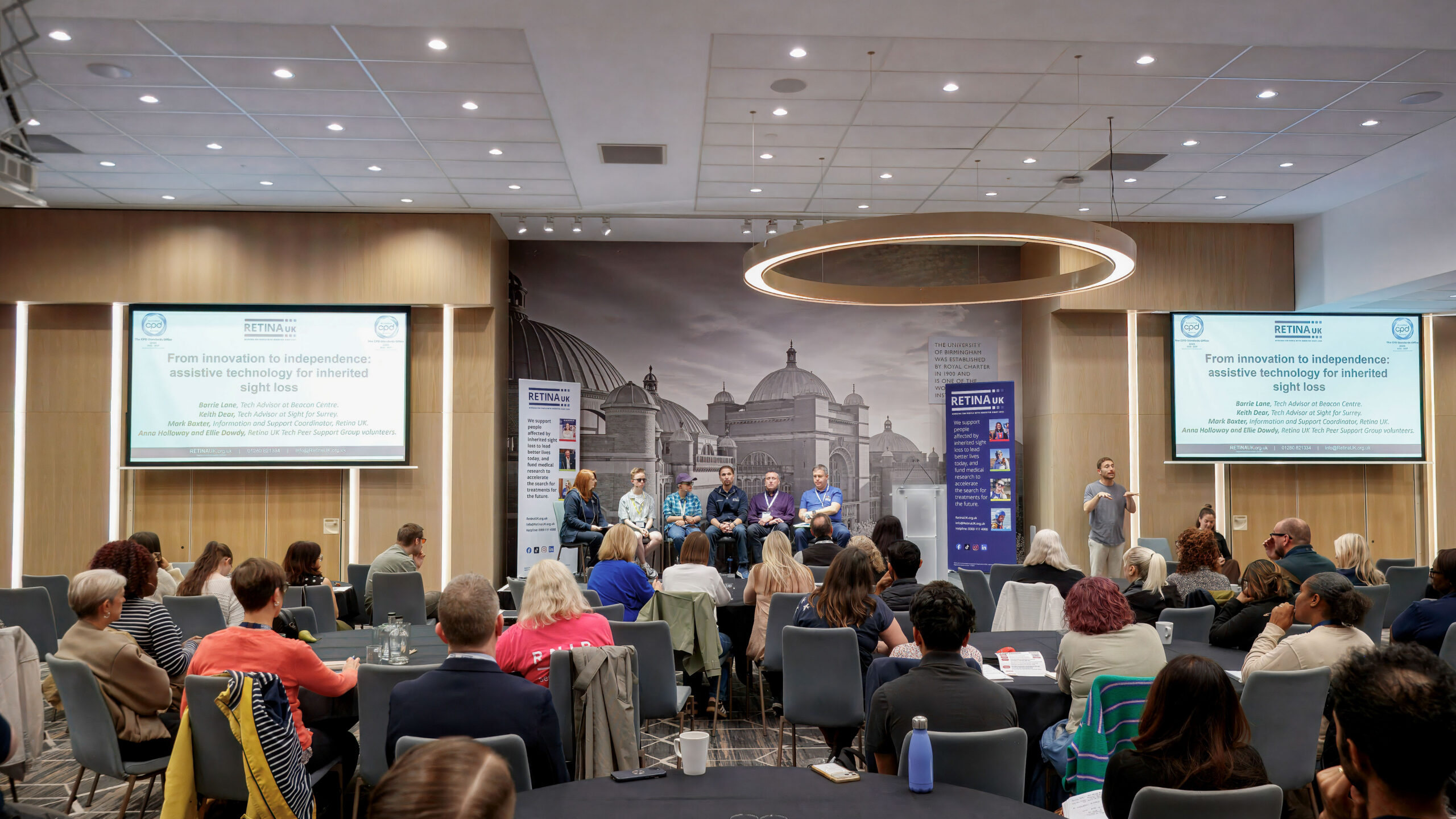 Picture shows the room in action at the Professionals' conference. There is a stage with 6 people sat speaking at a panel. Either side of them are a projector screen and in front are several round tables with delegates sat around them.