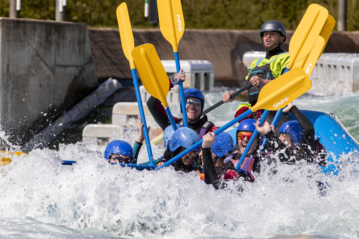 A blue boat with a group of people on board white water rafing. The yellow oars are raised.