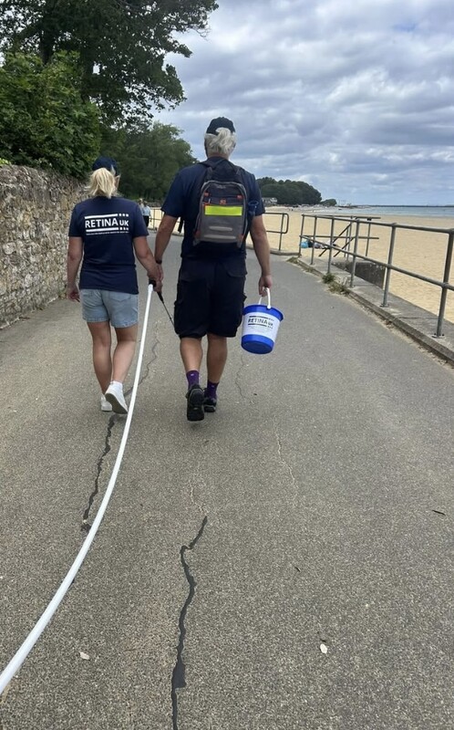 Two people seen from behind. One is wearing a Retina UK tshirt and the other is carrying a Retina UK collection bucket